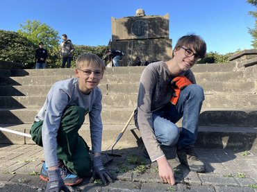 Auf dem Foto sind zwei Schüler zu sehen. Sie zupfen Unkraut, das zwischen den Pflastersteinen herauswächst. Beide Schüler schauen lächelnd in die Kamera.