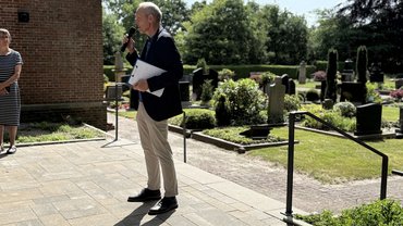 Der Lehrer hält in seiner Hand das Mikro und spricht zum Publikum. Mit der anderen Hand hält er sein Klemmbrett fest. Im Hintergrund ist wieder der Friedhof zu sehen.