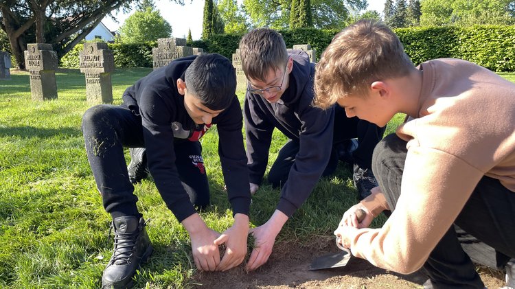 Drei Schüler befreien einen sandigen Gehweg von überschüssigem Rasen und Unkraut. Sie arbeiten kniend. Keiner schaut in die Kamera, sondern alle zum Boden. 
