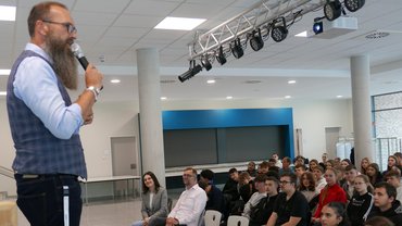 Links im Bild steht der Schulleiter. Er hält ein Mikrofon in seiner rechten Hand. Er schaut direkt zu den Schülerinnen und Schüler. Die Schüler sind rechts im Bild zu sehen. Sie sitzen alle gereiht und schauen zum Schulleiter.