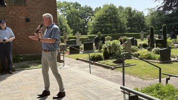 Der Bürgermeister spricht in Richtung des Publikums. Im Hintergrund ist der Friedhof zu sehen. Dort sind mehrere Grabsteine, ein Fußweg aus Schotter, über den man zu den Gräbern gelangt. 
