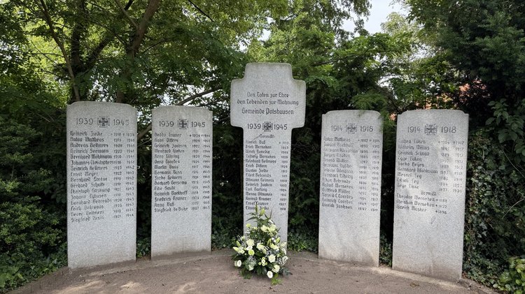 Zu sehen ist das Ehrenmal. Auf den Steintafeln stehen mehrere Namen. In der Mitte steht ein steinernes Kreuz. Vor dem Kreuz steht ein Blumengesteck.