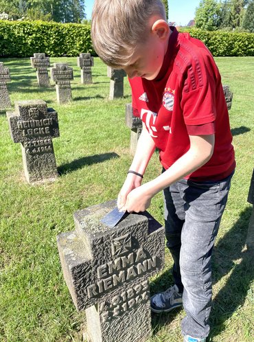 Auf dem Foto sieht man einen Grabstein eines deutschen Soldaten. Ein Schüler kratzt an der Oberfläche des Grabsteins Moos und andere Verschmutzungen ab. Im Hintergrund sieht mand weitere Kriegsgräber.
