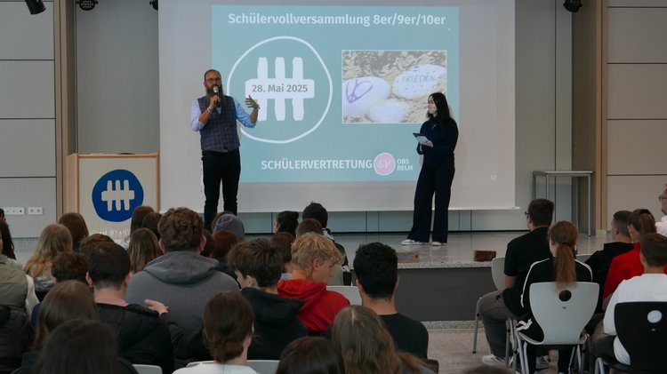 Zu sehen ist die Bühne und im Hintergrund eine Leinwand mit einer Präsentation. Links im Bild steht der Schulleiter und rechts die Schülersprecherin, die die Veranstaltung moderiert. Sie schaut zum Schulleiter. Vorne sind die Schüler aus dem Publikum zu sehen. Sie wurden von hinten fotografiert.