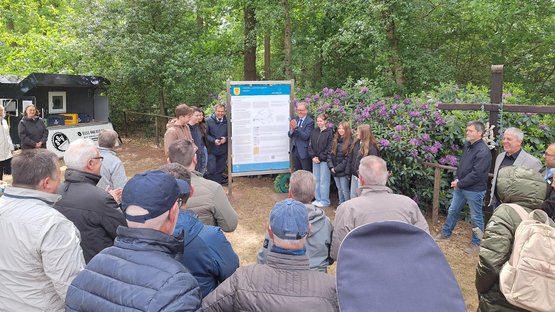 Im Vordergrund eine Gruppe von Menschen, mit dem Rücken zum Fotograf. Dahinter, in der Bildmitte, eine große Tafel mit Abbildungen und Texten. Links und rechts davon einige Menschen verschiedenen Alters.