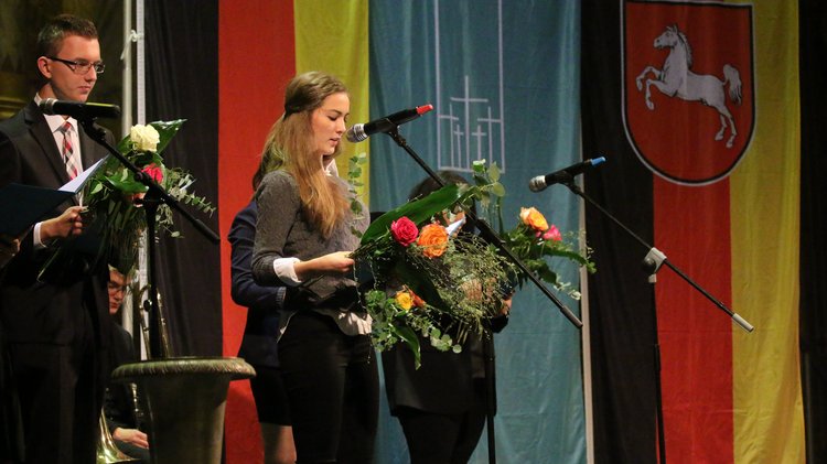 Links im Bild Jugendliche mit Blumensträußen. Ein Mädchen spricht in ein Mikrofon. Im Hintergrund von links nach rechts: Deutschlandflagge, Flagge des Volksbundes mit fünf Kreuzen und die Deutsche FLagge mit Niedersachsenwappen.