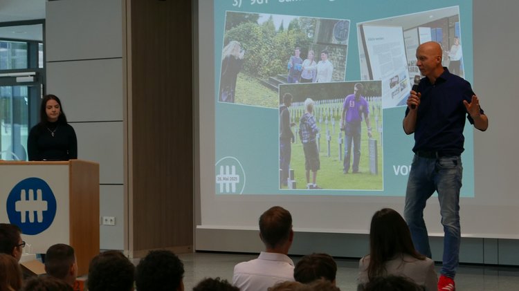 Links im Bild hinter dem Rednerpult steht die Schülersprecherin. Sie Schaut nach rechts zum Lehrer. Der Lehrer hält in seiner linken Hand das Mikrofon. Er spricht direkt zu den Schülern im Publikum.