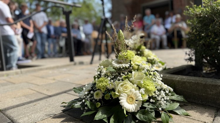 Im Vordergrund ist ein Blumengesteck. Die Blumen sind weiß. Im Hintergrund sieht man das Publikum, jedoch unscharf.