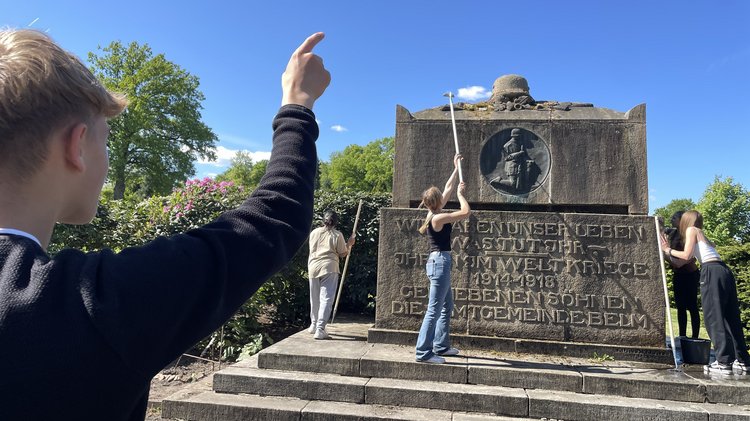 Auf dem Foto sieht man ein Gedenkstein, der an die gefallenen Soldaten erinnern soll. Rundum den Gedenkstein stehen Schülerinnen. Sie schrubben die Verschmutzungen vom Stein. Im Vordergrund des Bildes sieht man einen Schüler, der mit seinem Finger in ihre Richtung zeigt. 