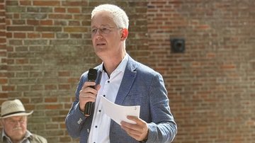Der Landrat hält eine Rede. Er schaut das Publikum dabei ein. Im Hintegrund sitzt ein Zuschauer. 