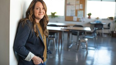 Blick in ein Klassenzimmer. Links im Bild eine Frau mit langen braunen Haaren. Sie lehnt an einer Wand und lächelt in die Kamera.