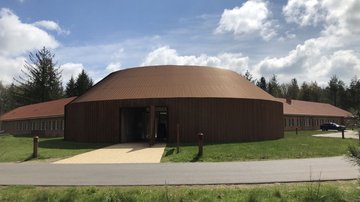 Mittig eine rundes Gebäude mit einer Eingangstür. Links und rechts hinten gegen weitere Gebäudeteile ab. 