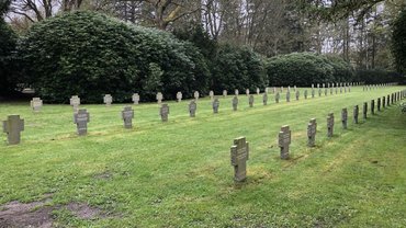 Auf einer Rasenfläche drei Reihen von Grabkreuzen. Im Hintergrund Büsche und Bäume.