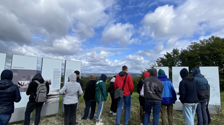 Menschegruppe vor Ausstellungstafeln die auf einem Hügel instaliert sind. Sie stehen auf einer Rasenfläche, über ihnen Wolkenformationen.