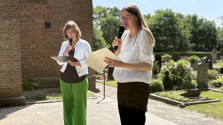 Zwei Personen halten eine Rede. Beide halten in ihrer Hand ein Mikrofon. Sie sprechen abwechelnd. Im Hintegrund ist der Friedhof. Es sind einige Grabsteine zu sehen.
